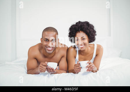 Heureux couple lying on bed tout en tenant tasses Banque D'Images