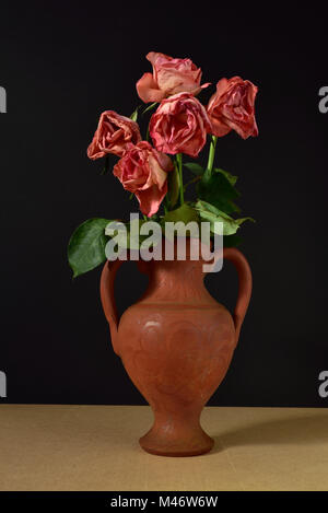 Cinq roses roses mourir dans un vase en terre cuite sur un fond noir. Banque D'Images