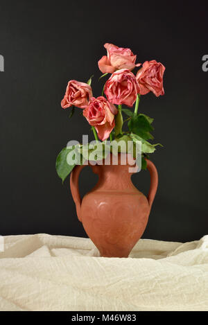 Cinq roses roses mourir dans un vase en terre cuite sur un fond noir. Banque D'Images