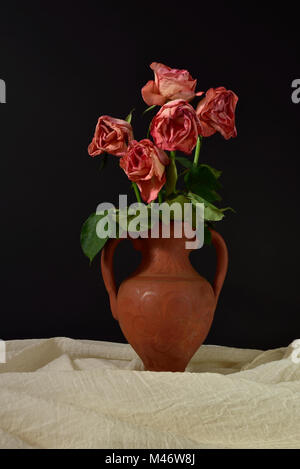 Cinq roses roses mourir dans un vase en terre cuite sur un fond noir. Banque D'Images
