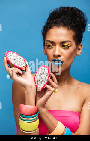 Photo de femme afro fascinant avec des cosmétiques sur le visage à la dragon fruit juteux sur réduit de moitié dans les deux mains isolated over Blue Ash Banque D'Images