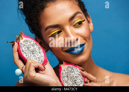 Portrait de femme mulâtre incroyable avec un miroir lumineux bénéficiant de pitaya exotique fruits coupés en deux et à côté isolé en studio sur blue backgroun Banque D'Images