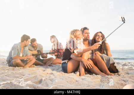 Smiling friends sitting on sand chanter et prendre des autoportraits Banque D'Images