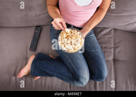 View of Woman Sitting on Sofa Eating Popcorn à côté de chez vous à distance Banque D'Images