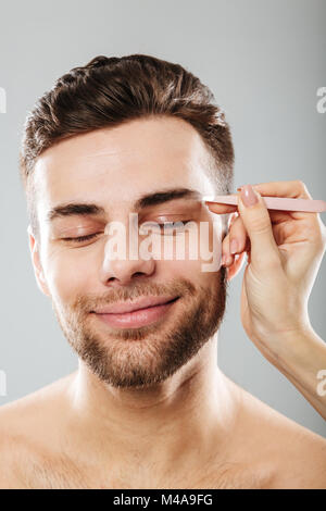 Portrait d'homme aux yeux clos détendue tout en souriant la main femme plumant ses sourcils avec des brucelles, isolé sur fond gris Banque D'Images