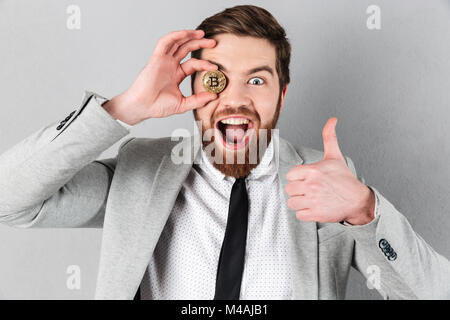 Close up d'un homme gai dressed in suit holding à bitcoin son visage sur fond gris isolé Banque D'Images