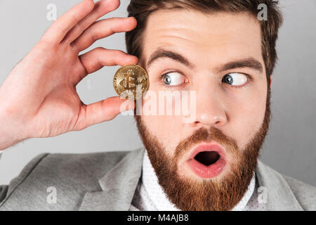 Close up of a businessman curieux dressed in suit holding à bitcoin son visage sur fond gris isolé Banque D'Images