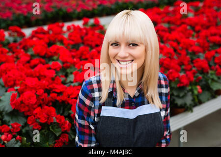 Charmante femme gaie permanent jardinier contre de grandes plantations de fleurs bégonia rouge, looking at camera. Banque D'Images
