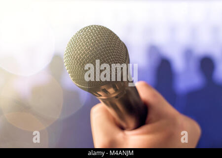 La prise de parole en public et de donner expression concept. Microphone main tenant devant une foule de gens silhouette avec des reflets et de la lumière du soleil flou de fuite. Banque D'Images