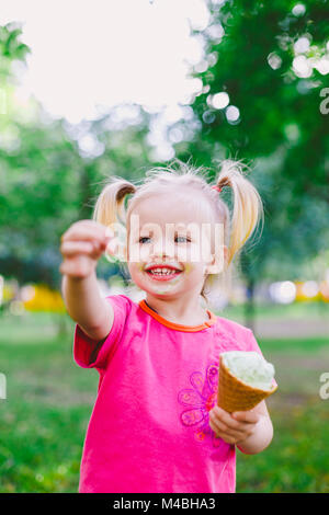 Peu drôle girl blonde bleu doux de manger de la crème glacée dans un cornet gaufré cup sur un arrière-plan vert de l'été dans le parc. enduit son visage et les joues et rit. Vêtus de vêtements lumineux et élégant Banque D'Images