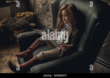 Fille de l'enfant dans l'image de Sherlock Holmes est assis dans un fauteuil et lit journal sur fond de murs en briques anciennes et minable et parquet. Banque D'Images