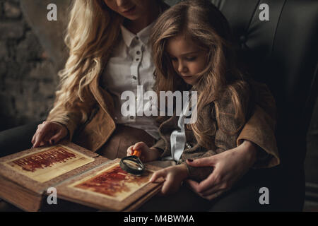 Enfant fille avec femme dans l'image de Sherlock Holmes est assis dans un fauteuil et l'air vieux photoalbum avec loupe sur fond de murs en briques anciennes. Banque D'Images