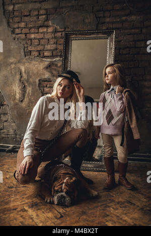 Enfant fille avec femme dans l'image de Sherlock Holmes se trouve à côté de bulldog Anglais sur fond de murs en briques anciennes, miroir et minable et parquet. Banque D'Images