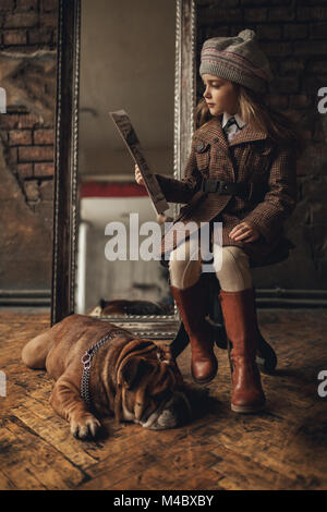 Fille de l'enfant dans l'image de Sherlock Holmes s'assoit à côté de bulldog Anglais et lit journal sur fond de murs en briques anciennes, miroir et minable en fl Banque D'Images