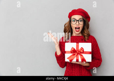 Portrait d'une femme surpris vêtu de rouge chandail et lunettes holding gift box isolé sur fond de mur gris Banque D'Images