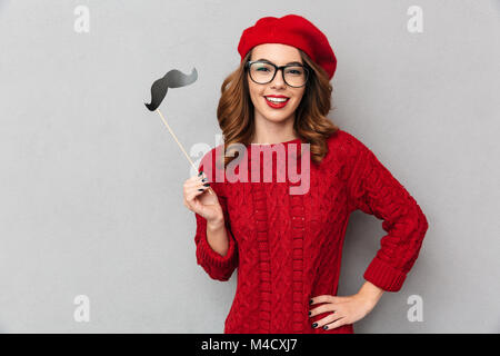 Portrait of a smiling woman dressed in chandail rouge et les lunettes holding fake moustache et à la caméra au fond de mur gris plus isolés Banque D'Images