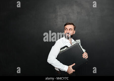 Portrait of happy riche businessman carrying briefcase full of dollar bills et regarde en arrière sur fond gris foncé isolés Banque D'Images