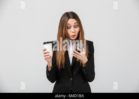 Photo de jeune chat businesslady choqué en smartphone et holding tasse de café plus graywall isolés Banque D'Images