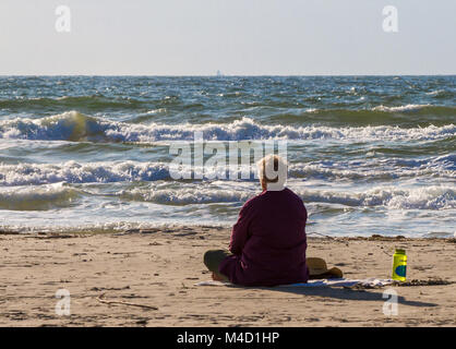 Un Américain Senior citizen femme assise sur la plage et regarde l'océan dans la lumière du soleil tôt le matin. Banque D'Images