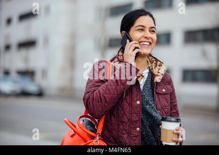 Femme marche dans la ville avec une tasse de café tout en parlant au téléphone. Banque D'Images