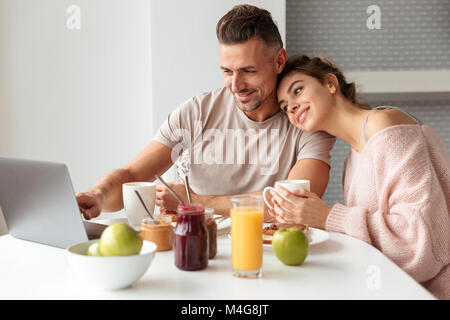 Portrait of a happy couple aimant prendre le petit déjeuner alors qu'il était assis à la table dans une cuisine à la maison et à l'aide d'un ordinateur portable Banque D'Images