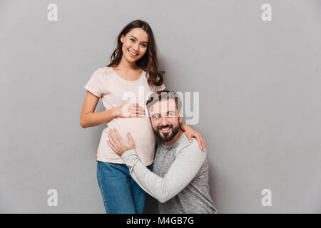 Portrait of a smiling young man listening to his pregnant wife's belly assis sur fond gris Banque D'Images