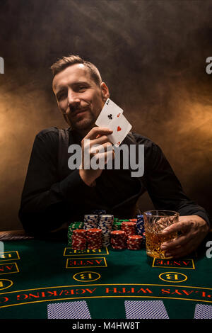 Beau joueur de poker avec deux as en main et jetons de poker assis à table dans une pièce sombre pleine de fumée de cigarette. Cartes de visite, plaquettes, de whisky, de cigare Banque D'Images