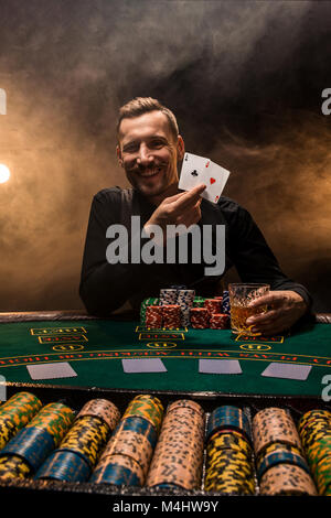 Beau joueur de poker avec deux as en main et jetons de poker assis à table dans une pièce sombre pleine de fumée de cigarette. Cartes de visite, plaquettes, de whisky, de cigare Banque D'Images