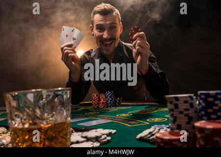 Beau joueur de poker avec deux as en main et jetons de poker assis à table dans une pièce sombre pleine de fumée de cigarette. Cartes de visite, plaquettes, de whisky, de cigare Banque D'Images