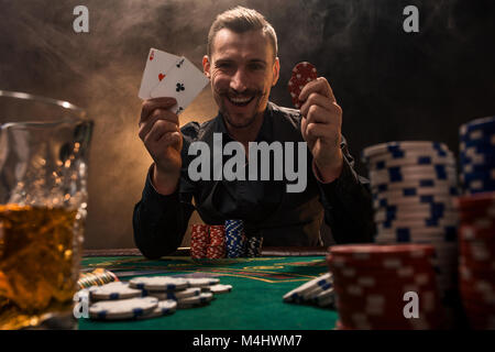 Beau joueur de poker avec deux as en main et jetons de poker assis à table dans une pièce sombre pleine de fumée de cigarette. Cartes de visite, plaquettes, de whisky, de cigare Banque D'Images