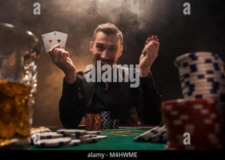 Beau joueur de poker avec deux as en main et jetons de poker assis à table dans une pièce sombre pleine de fumée de cigarette. Cartes de visite, plaquettes, de whisky, de cigare Banque D'Images