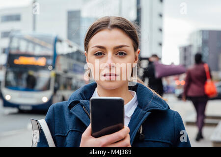 Portrait d'une jeune femme avec un téléphone intelligent. Elle se tient dans la ville et est à la recherche de l'appareil photo. Banque D'Images