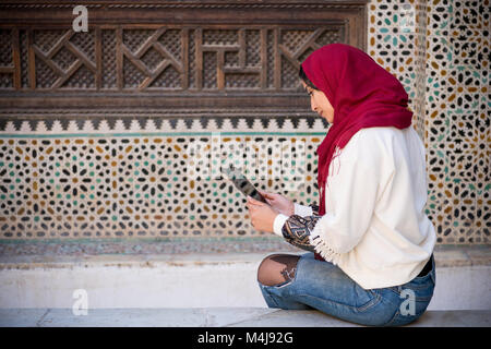 Femme musulmane travaillant sur le comprimé dans des vêtements traditionnels avec foulard rouge sur sa tête à côté d'un mur traditionnel marocain arabesque Banque D'Images