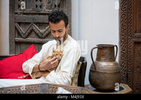 Jeune homme en costume traditionnel musulman tenant un chat jaune devant un mur décoré Banque D'Images