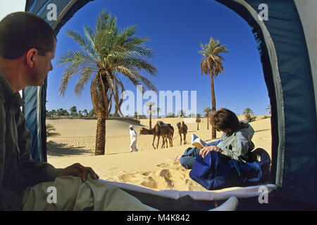 L'Algérie. Près de Timimoun. La mer de sable de l'Ouest. Grand Erg occidental. Désert du Sahara. Le camping. Tente, touristes, couple, homme, femme, lecture, palmiers, camel Banque D'Images