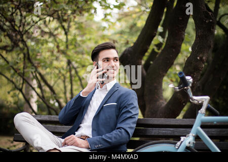 Young businessman talking on smartphone à l'extérieur dans un parc, assis sur un banc avec son vélo à côté de lui. Banque D'Images