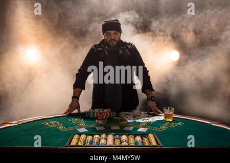 Jouer au poker joueur de sexe masculin, l'intensité de couleur sombre fumée. Un grand homme barbu avec un verre de whisky dans les mains s'élève au-dessus de la table de poker Banque D'Images