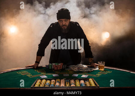 Jouer au poker joueur de sexe masculin, l'intensité de couleur sombre fumée. Un grand homme barbu avec un verre de whisky dans les mains s'élève au-dessus de la table de poker Banque D'Images