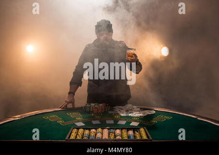 Jouer au poker joueur de sexe masculin, l'intensité de couleur sombre fumée. Un grand homme barbu avec un verre de whisky dans les mains s'élève au-dessus de la table de poker Banque D'Images