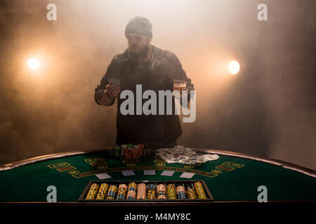 Jouer au poker joueur de sexe masculin, l'intensité de couleur sombre fumée. Un grand homme barbu avec un verre de whisky dans les mains s'élève au-dessus de la table de poker Banque D'Images