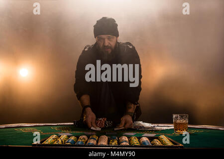 Jouer au poker joueur de sexe masculin, l'intensité de couleur sombre fumée. Un grand homme barbu avec un verre de whisky dans les mains s'élève au-dessus de la table de poker Banque D'Images