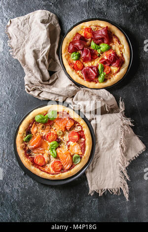 Deux pizza traditionnelle avec bresaola, saumon fumé, fromage, tomates et basilic servi sur les plaques noires avec dessus textile texture background noir. Haut Banque D'Images