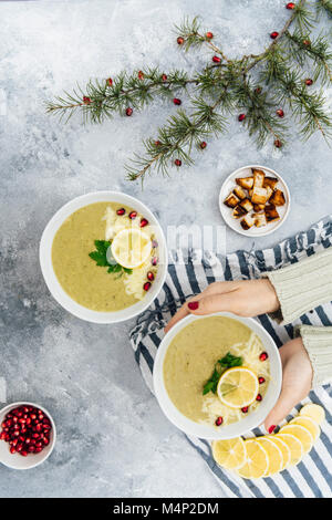 Une femme tenant un bol de soupe au brocoli fromage photographié en vue de dessus. Les arilles de grenade, les tranches de citron, croûtons et pine tree branches accompagner. Banque D'Images