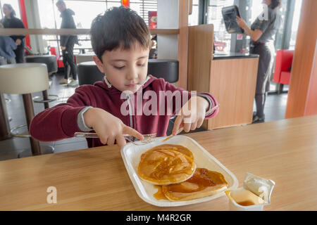 Comme un plaisir, un jeune enfant bénéficie des Crêpes avec sirop pour le petit-déjeuner dans un restaurant McDonalds. Banque D'Images