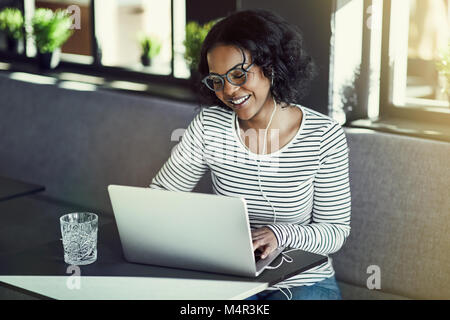 Smiling young African woman wearing earphones et en utilisant son ordinateur portable afin de discuter en ligne alors qu'il était assis seul à une table Banque D'Images