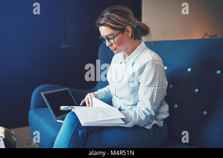 Young businesswoman reading à travers des documents et à l'aide d'un ordinateur portable tout en étant assis sur un canapé à la maison Banque D'Images