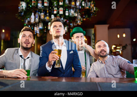 Les barbus assis au comptoir du bar et regarder des match de rugby avec concentration, red-haired homme portant costume lutin debout derrière eux Banque D'Images