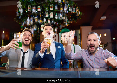 Groupe d'hommes barbus célébrant la victoire de leur équipe de football préférée tout en étant assis au comptoir du bar avec un verre de bière dans la main et regarder des match Banque D'Images