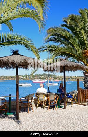 Un couple de détente à la terrasse d'un café le long de la promenade avec vue sur des yachts amarrés dans l'estuaire, Alvor, Algarve, Portugal, Europe. Banque D'Images