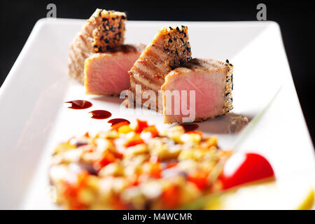 Sur une assiette blanche, des morceaux de thon dans le sésame avec gouttes de sauce rouge garnir de riz légumes. Fond noir Banque D'Images